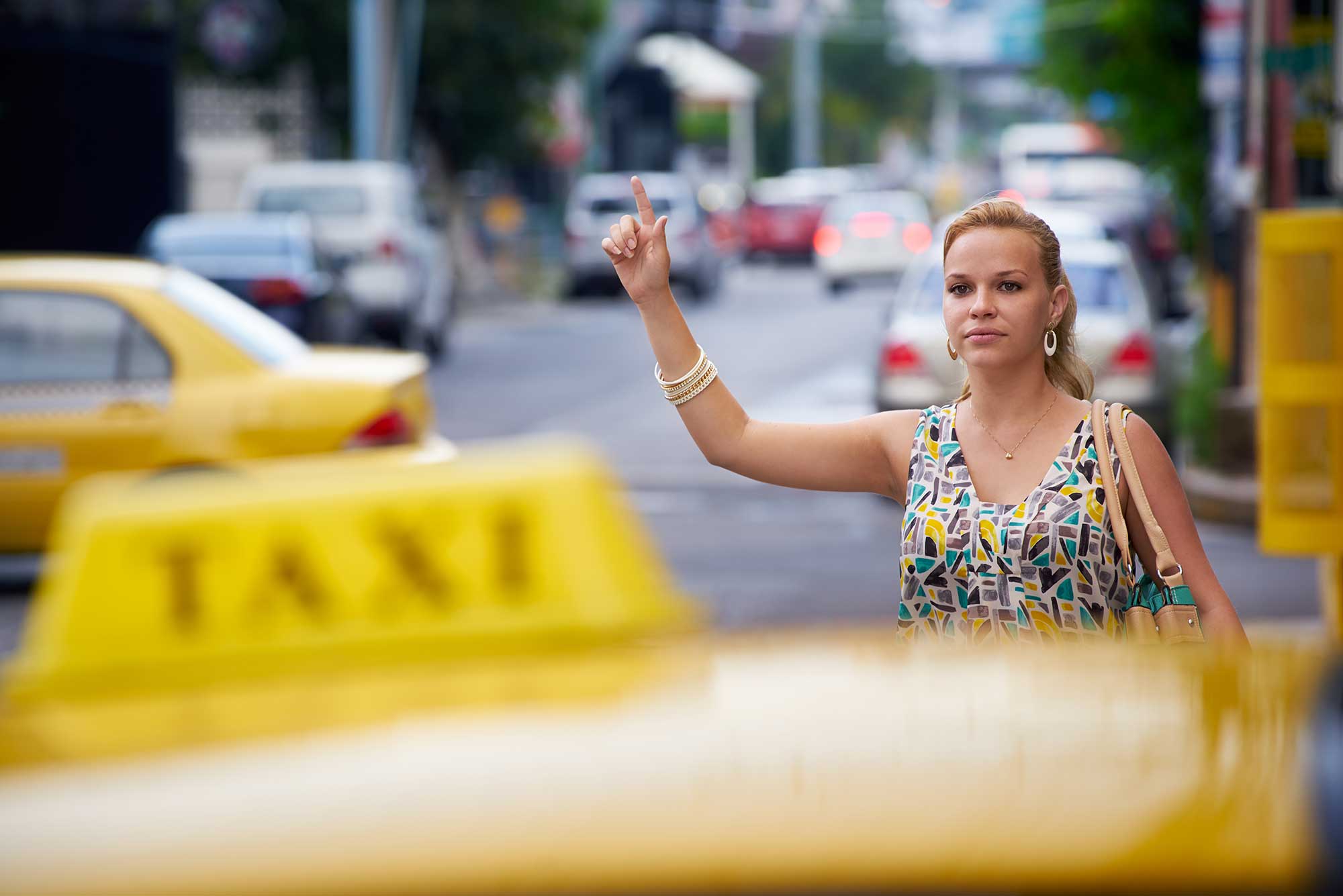 A woman stands on the street, raising her hand to hail a taxi amidst busy traffic. She is wearing a colorful dress and appears focused on getting a ride.