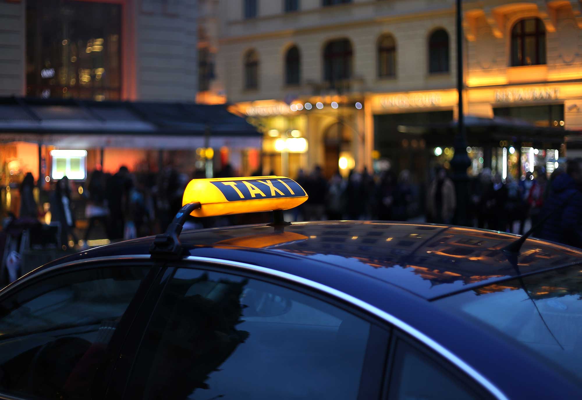 A taxi with its illuminated sign is parked in an urban area at night, surrounded by a blurred crowd and illuminated buildings.