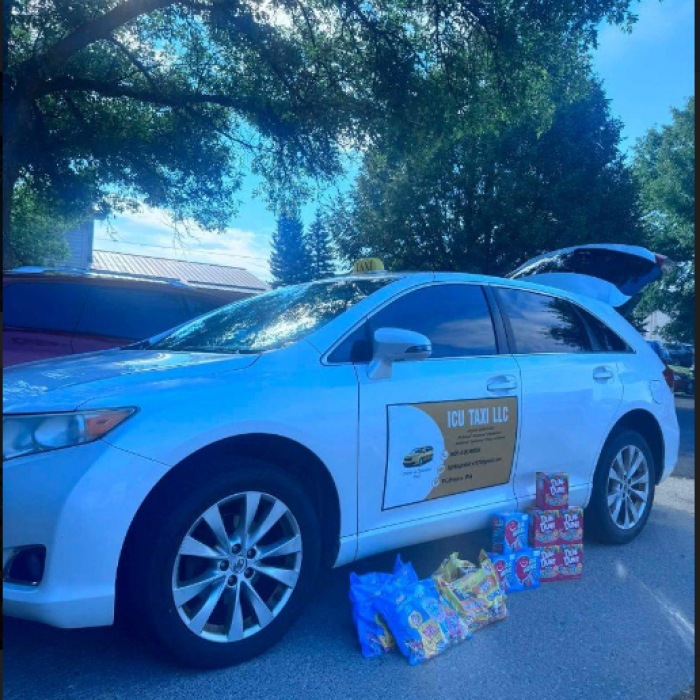 Blue sedan parked beneath leafy trees, displaying a charity sign and surrounded by colorful bags and boxes of donated food, creating a generous mood.