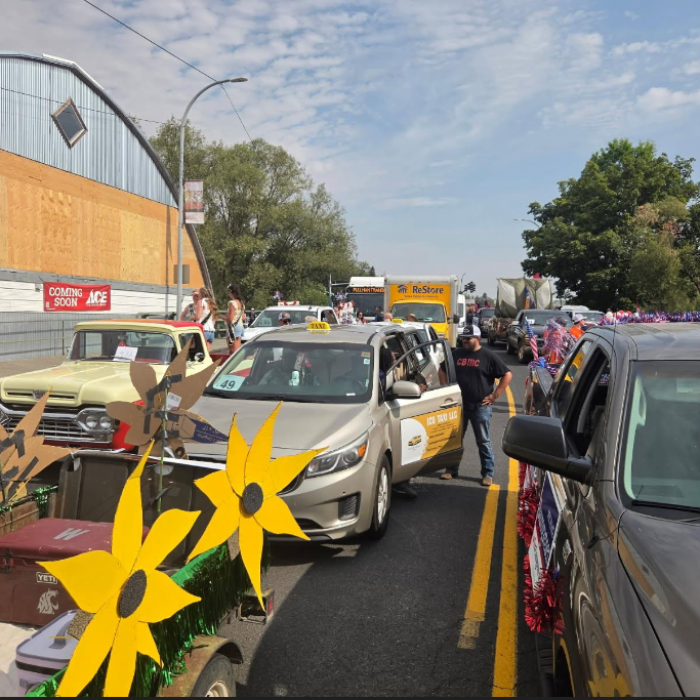 Colorful street parade with decorated cars, giant yellow flowers, cheering crowds, and a sunny urban road lined with trees and buildings.