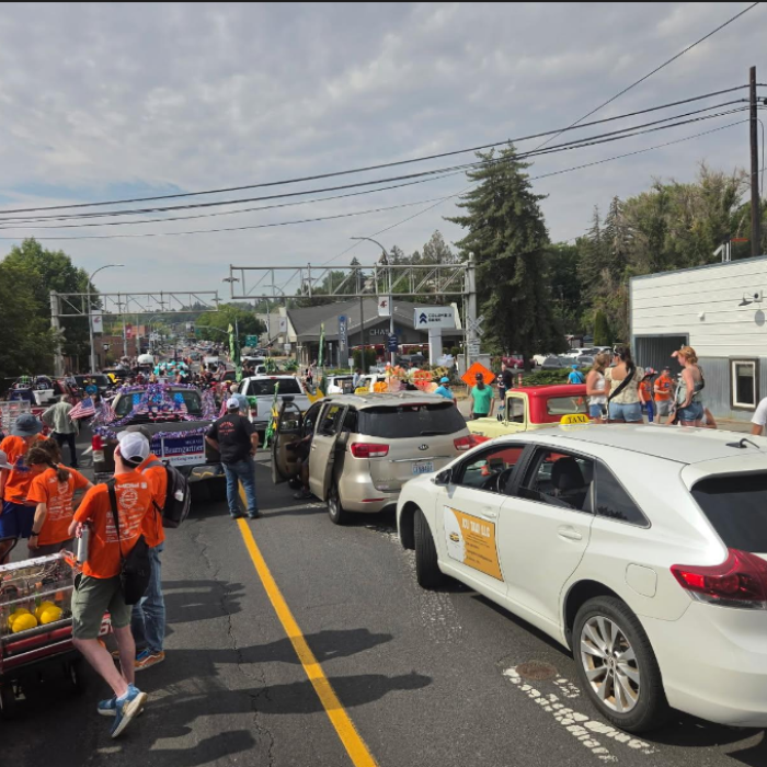 Crowded street festival with orange-shirted volunteers, decorated cars, and cheering spectators beneath cloudy skies and overhead wires.