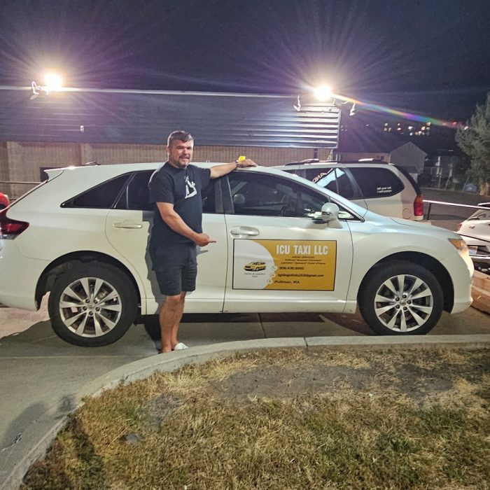 Man standing beside a white taxi at night in a parking lot, with bright lights, nearby vehicles, trees, and dry grass visible.